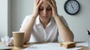 Persona seduta alla scrivania con le mani alla testa, segni di stress e affaticamento davanti a documenti e un caffè