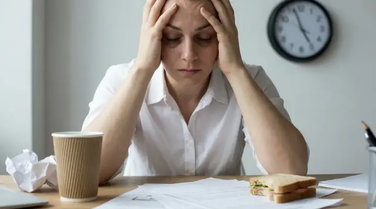 Persona seduta alla scrivania con le mani alla testa, segni di stress e affaticamento davanti a documenti e un caffè