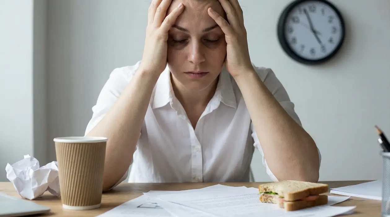 Persona seduta alla scrivania con le mani alla testa, segni di stress e affaticamento davanti a documenti e un caffè