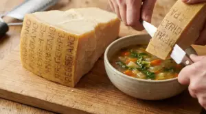 Mani che aggiungono la scorza di Parmigiano Reggiano a una zuppa di verdure su tagliere di legno