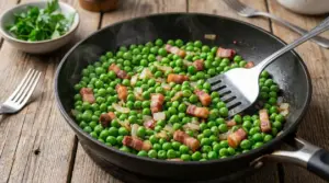 Padella con piselli in cottura con cipolla e pancetta, mescolati con una spatola su tavolo di legno